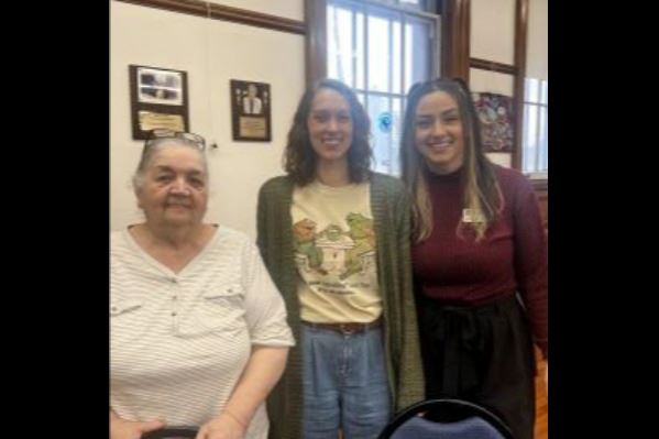 A senior stands with her hands on the back of a chair, along with two smiling young women.