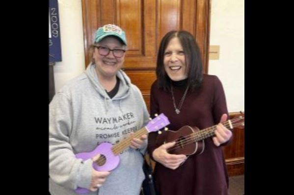 Two women in jeans play a purple ukulele and a brown ukulele.