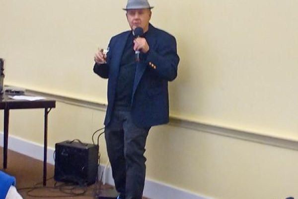 A man in a fedora holds a microphone in one hand and a martini in the other.