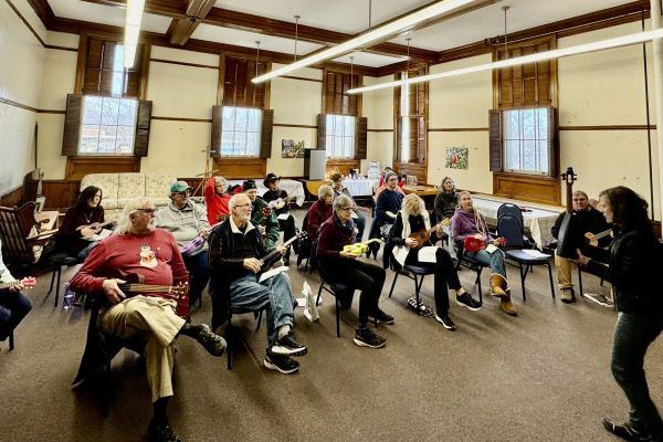 Over a dozen people seated in a classroom hold or strum ukuleles.